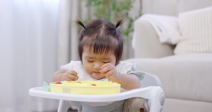 Asian toddler girl sitting on feeding chair picks strawberry piece from yellow plate, stares at fruit curiously, soft natural light from window, quiet morning mood, self feeding habit, child learning