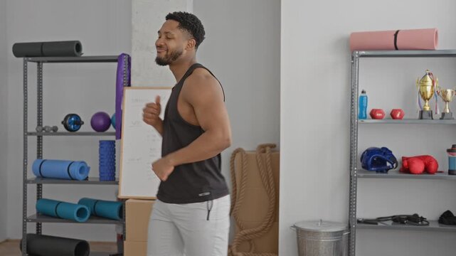 Man with bare arms shadowboxing and smiling, fists raised near yoga mats, foam rollers, dumbbells, boxing gloves and a whiteboard in a training studio; joy confidence focus.