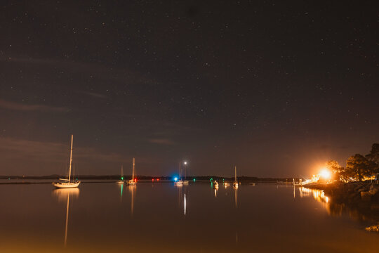 Sailboats at anchor under a starry night sky with reflections on calm water