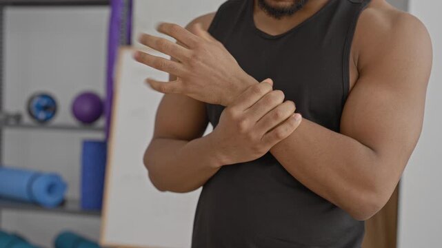 Man massaging sore wrist and forearm after training, wearing black tank top with visible muscles in studio; recovery.