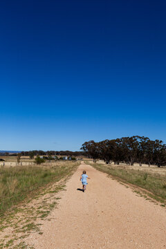 Little farm girl under blue summer sky running down gravel road through paddocks
