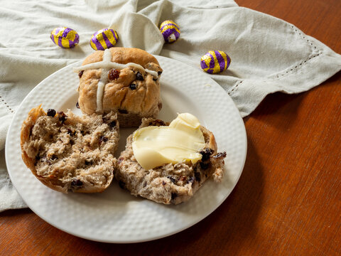 Two hot cross buns, one with butter, on white plate with linen napkin and foil eggs