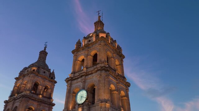 Cathedral Basilica of the Immaculate Conception in Durango, Mexico glows warmly at sunset. Twin baroque bell towers with ornate stone carvings rise against a blue twilight sky with pink wispy clouds.