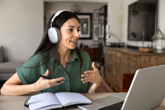 Woman in headphones speaks during video call, remote coaching at home office sits at table with laptop and notebook in living room. Online coaching, tutoring, consulting services, videoconferencing
