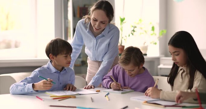 Young adult woman teacher assisting to diverse children drawing and coloring, developing skills during creative group art class activity in classroom. Primary school education, schooling concepts