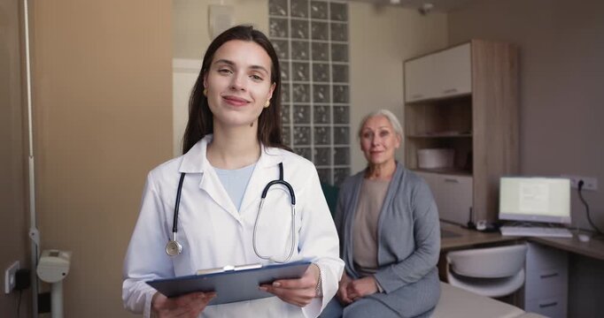Smiling female doctor holds clipboard, posing for portrait with senior woman sits on examination table in medical hospital office, representing healthcare services, medicine, professional patient care