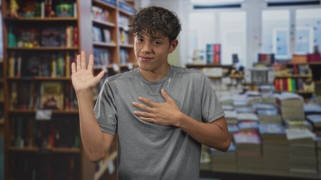 Teenage boy with palm raised and hand on chest wearing gray shirt and silver chain in a bookstore building; sincerity promise.