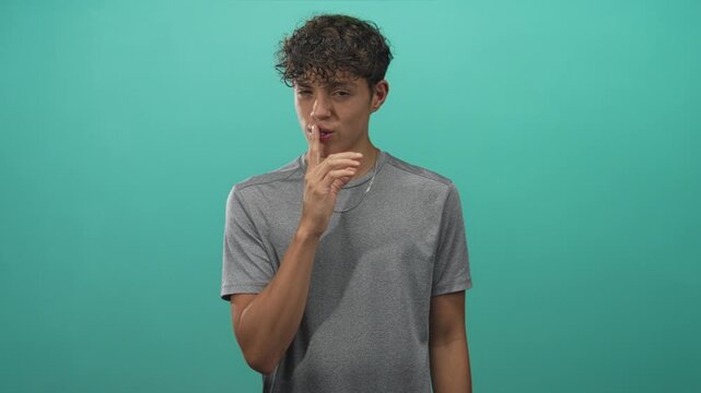 Man with finger to lips for silence in studio, smiling, wearing gray tshirt and silver chain; playful secret.