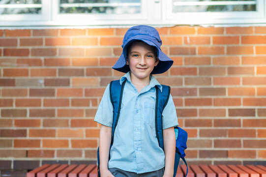 Boy in a blue school uniform and hat poses outside school with a backpack on a sunny day