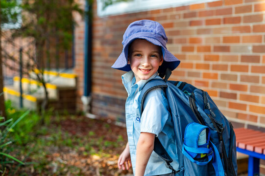 A young boy in a school uniform stands outside a primary school wearing a hat and smiling