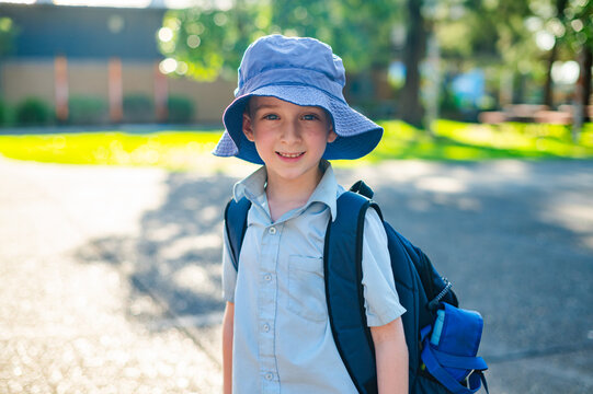 A boy wearing a school uniform and hat stands outside with a backpack, smiling on a sunny day