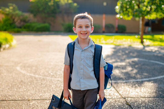 A young boy in a school uniform stands in a playground holding his school bag, smiling