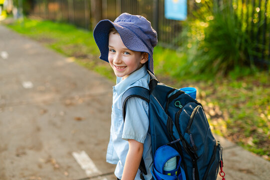 A boy in a school uniform carries a backpack and water bottle as he walks home, smiling