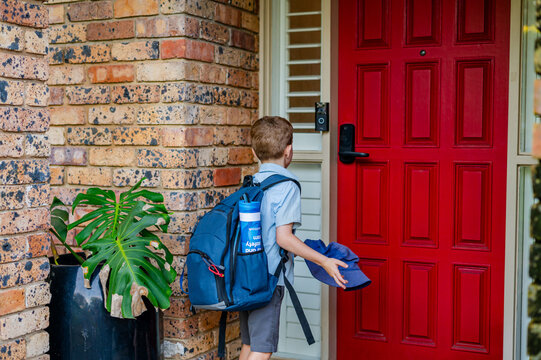 Boy stands at a red door holding a hat and wearing a backpack while arriving home after school