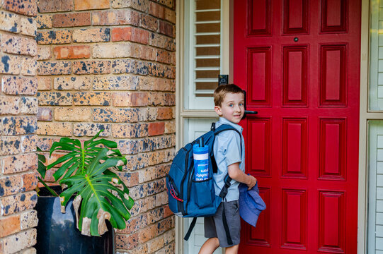 A boy with a backpack stands near a red door, ready to go inside after school