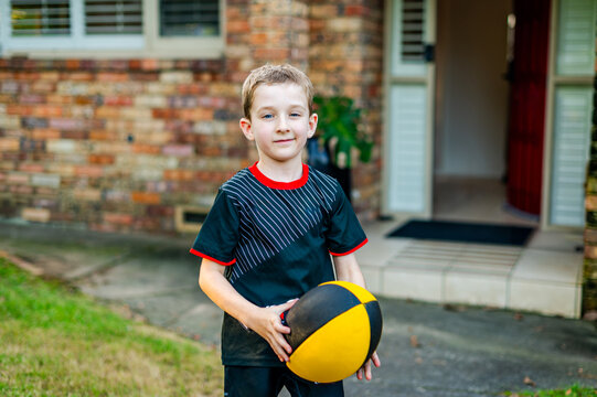 A young boy stands in front of his home holding a rugby ball, eager to start playing the sport