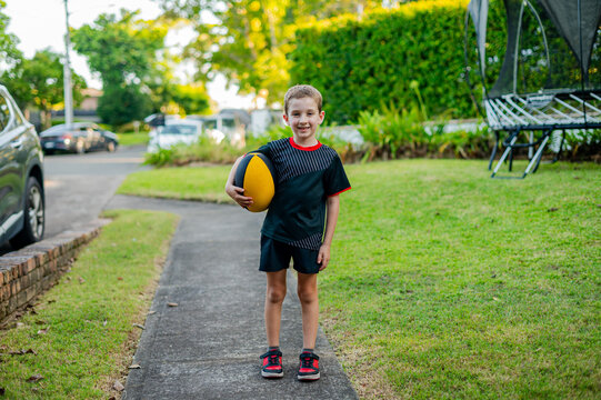 Boy smiles while on a path in his rugby uniform, holding a rugby ball, preparing to play outside