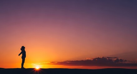 Fototapeta premium A person standing on a hillside at sunset, silhouetted against the vibrant colors of the sky.