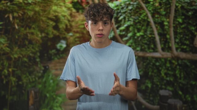 Teenage boy gesturing with hands while speaking in forest walkway with wooden railing and dense foliage; thoughtful reflection.