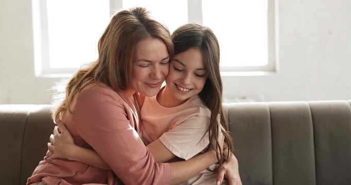 Affectionate Caucasian mother embracing her smiling child daughter sitting together on couch at home, showing support, expressing caress and love. Motherhood, family bonding and relationships concept