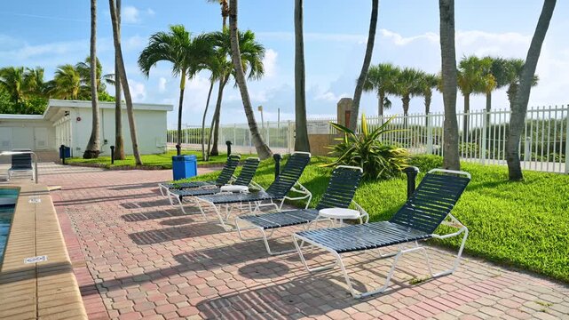 Sunbeds on paved resort area with white fence. Row of empty lounge chairs on a brick patio next to a swimming pool area with palm trees and a security fence.