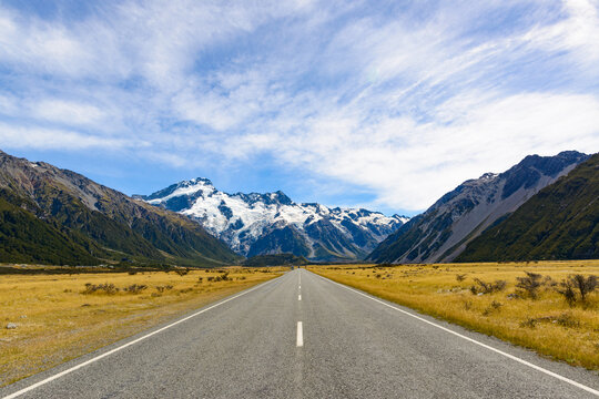 Low view of a road with line markings converging on a snow capped mountain with blue sky and clouds