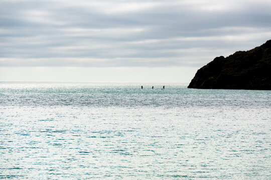 Minimalist image of three paddlers in the distance with grey green sea and grey sky and a headland