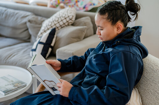 Teenage Samoan girl sitting comfortably on the couch reading a book in the living room