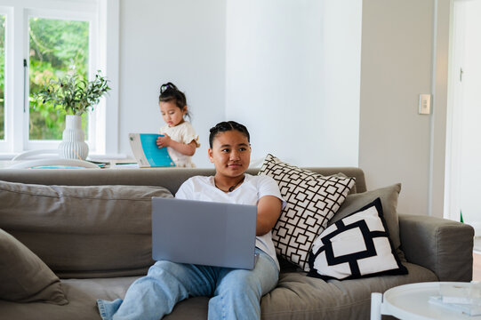 Teenage girl sitting in the couch with her laptop while younger sister opening a book behind her