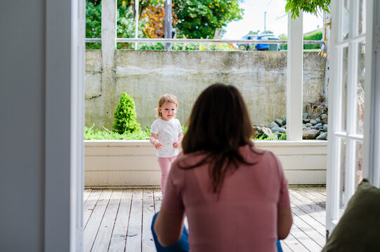 Toddler girl playing in the wooden deck outdoors while mum sits in the doorway