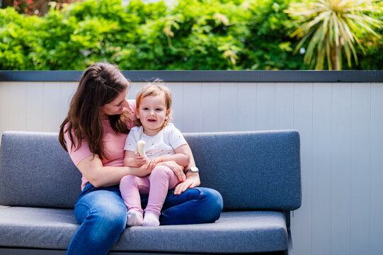 Italian mum sitting on grey couch with toddler girl on her lap holding a banana on the balcony