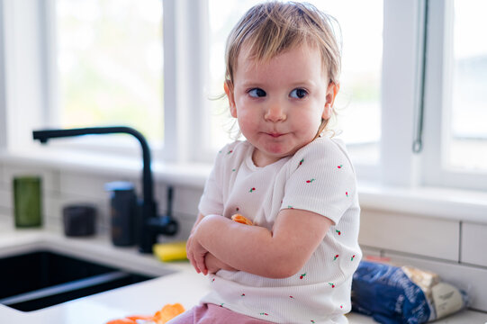 Toddler girl sitting on kitchen counter with arms folded across her chest to holding orange wedges