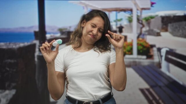 Woman holding contact lens case between fingers on a street promenade near the seaside, pinching tiny lens while squinting in bright sun; disgust.
