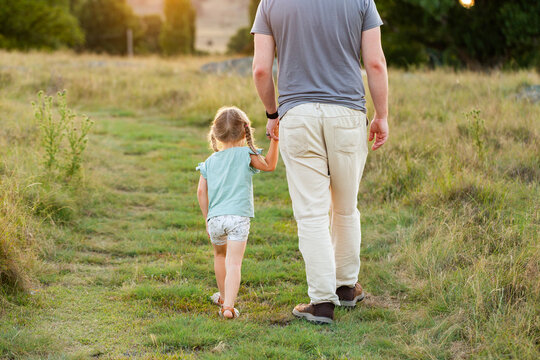 Dad on walk with young daughter walking away down grassy path in recreational area