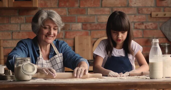 Loving grandmother and granddaughter flattening dough with rolling pins, cook together, baking pastry in kitchen. Home cooking, knowledge sharing, multigenerational family bonding and communication