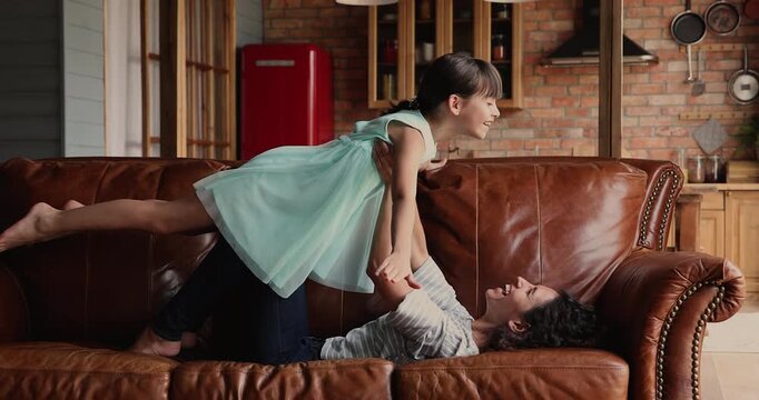 Mother lying on sofa, raising up her child, daughter imagining flying in air, playing at home, having fun, spending time together on weekend in living room. Family playtime, indoor activity with kid
