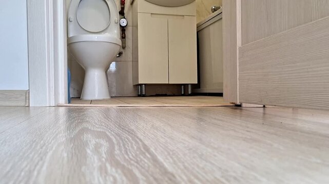 A low angle view of a bright, clean bathroom featuring a white ceramic toilet and a light-colored vanity cabinet. The shot highlights the smooth wood flooring and tiled bathroom area