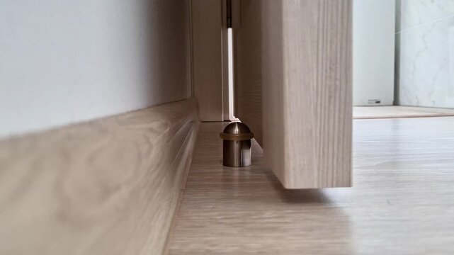 Metallic door stopper or magnetic holder securing door near wall corner on light wooden floor, preventing damage and highlighting functional detail of everyday household utility