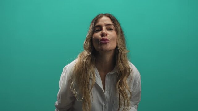 Woman puckering lips and blowing a kiss with face close to camera, holding a tablet in a green studio; playful greeting.