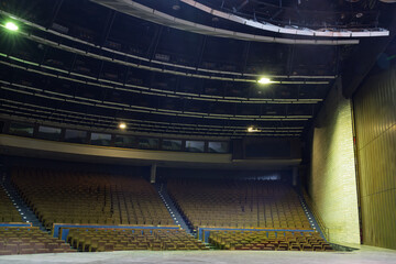 Dramatic stage view of a vast, empty abandoned concert hall with dark curved seating rows and balconies. Atmospheric dim lighting illuminates faded seats and a yellow brick wall in decaying auditorium © Aimur