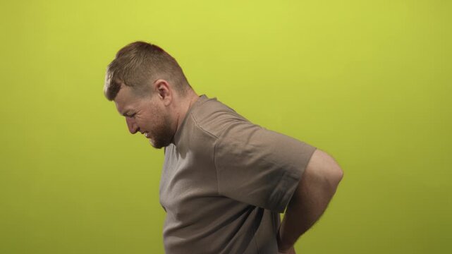 Young overweight man holding lower back and grimacing in lime green studio, profile view and casual posture; backache pain recovery.