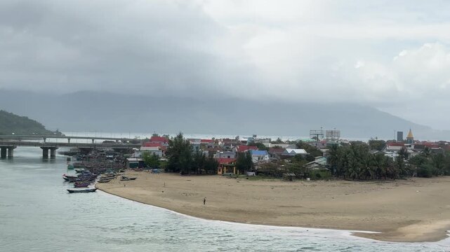 Train-window view of a sandy river mouth and coastal town in central Vietnam, with fishing boats, bridge, and stormy mountains beyond the shoreline.