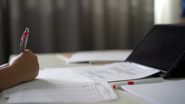 closeup hand of child learn from home write on paper or young kid girl student math calculate or doing homework with computer tablet on desk by study online class or people back to school learning