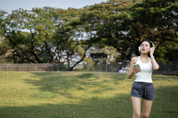 Fototapeta premium Happy Asian woman in fitness apparel with white headphones using smartphone in urban park. Young girl relaxing with music and wellness app, city lifestyle, workout break, and personal relax