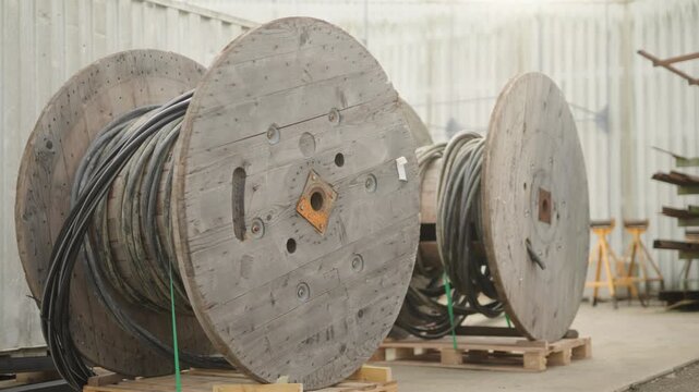 Static shot of large wooden cable reels with coiled industrial cables stored outdoors in an industrial or construction environment, in daylight, with no people visible.