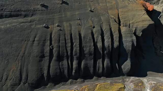 Aerial view of a dark eroded volcanic slope with vertical ridges, scattered boulders, and striking shadow patterns in a remote barren landscape.