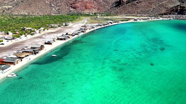4K drone flying over the water toward shore at Ballena Pel&eacute;chada near Muleg&eacute;, passing beachfront shacks, cars, and RVs along the desert coastline.