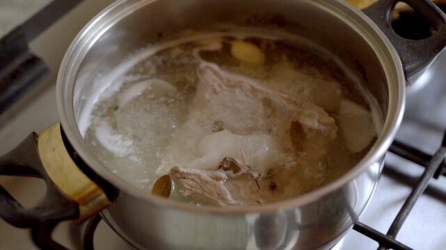 Closeup of hands stirring boiling chicken and meat in pot on gas stove with bubbling broth showing cooking process and food preparation in kitchen