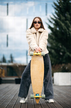 Young woman in stylish wear posing on roof with skateboard