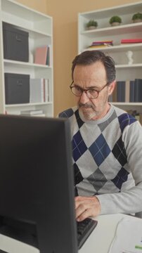 Man middle age hispanic wearing glasses holding phone to ear and typing on computer keyboard while reviewing documents at desk in building; concern focus.